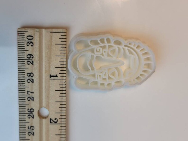 May include: A white plastic cookie cutter in the shape of a stylized face, approximately 2.5 inches long. The design includes facial features and decorative elements around the face. A wooden ruler is next to the cookie cutter.