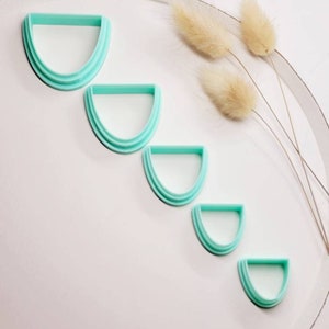 Puede incluir: Conjunto de cinco cortadores de galletas de plástico turquesa en forma de semicírculos, dispuestos en orden descendente de tamaño.