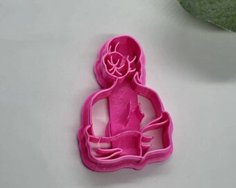 May include: Pink plastic cookie cutter shaped like a person sitting in a yoga pose with their legs crossed.