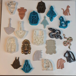 May include: A collection of various-shaped cookie cutters, including vases, pharaohs, cats, and snakes. The cutters are in shades of brown, blue, gray, and white. The shapes are detailed and suitable for creating themed cookies or decorative elements.