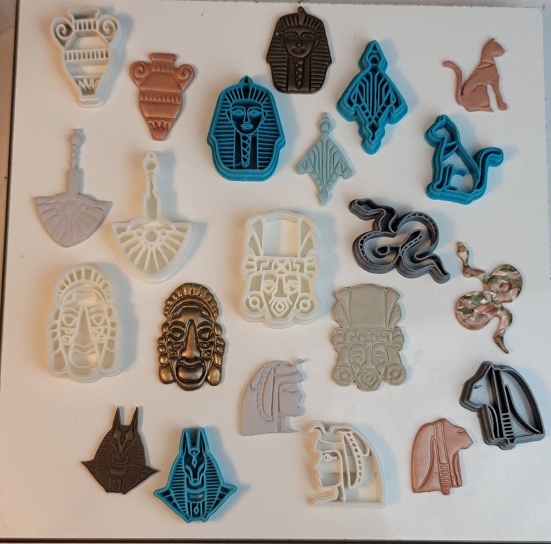 May include: A collection of various-shaped cookie cutters, including vases, pharaohs, cats, and snakes. The cutters are in shades of brown, blue, gray, and white. The shapes are detailed and suitable for creating themed cookies or decorative elements.