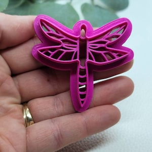 Puede incluir: Un cortador de galletas de libélula de plástico rosa. El cortador tiene un diseño detallado con un cuerpo delgado y delicado y alas.