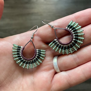 May include: Handmade macramé earrings with a fan design. The earrings feature a teardrop-shaped silver hook and a half-circle macramé design in shades of green, black, and burgundy. The earrings are held in a hand.