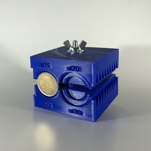 Op de afbeelding: Blauwe plastic muntpers met een zilveren schroef en een 2 euro munt erin. De pers heeft markeringen voor verschillende muntgroottes: 15, 20, 25 en 30.