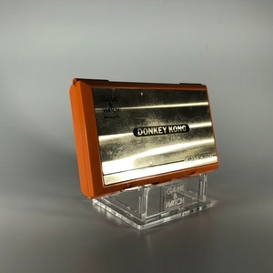 以下が含まれることがあります： オレンジとゴールドの任天堂ゲーム＆ウォッチ ドンキーコングの携帯ゲーム機。ゲームは透明なアクリルスタンドに展示されています。コンソールには「DONKEY KONG」と「MULTI SCREEN」の文字があります。