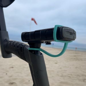 Può includere: Un metal detector nero con un attacco per manico di colore turchese. Il detector si trova su una spiaggia sabbiosa.