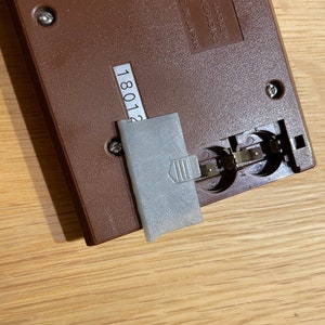 May include: Close-up of the back of a brown handheld game console with a gray battery cover. The console has a serial number "18012" printed on the back. The console has a label with the text "NO. PR-21 DCOV=0.0002W RY: LR44 or SR43) x2 Tende 1981 PEND.. MADE IN JAPAN".