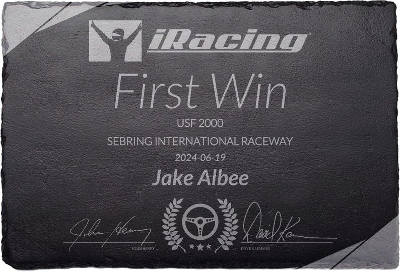 Puede incluir: Placa de pizarra negra grabada con el logo "iRacing" y "First Win". Detalles: "USF 2000", "Sebring International Raceway", fecha y nombre. Presenta un volante y dise&ntilde;o de corona de laurel.