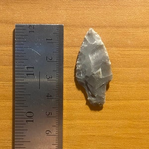 May include: A grey stone arrowhead measuring approximately 3.8 cm in length, next to a ruler.
