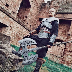May include: A person wearing a black tunic and leather armour with a fur collar holds a large, ornate axe with a silver blade and intricate carvings. The person is standing in front of a stone wall with an arched doorway.