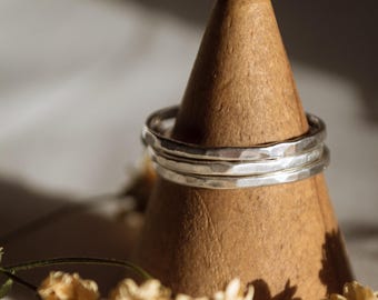 Könnte beinhalten: Drei silberne, gehämmerte Ringe, die auf einem Holzständer gestapelt sind. Die Ringe sind schlicht und zart, mit einer leicht unregelmäßigen Form. Der Ständer ist hellbraun und hat eine Kegelform.