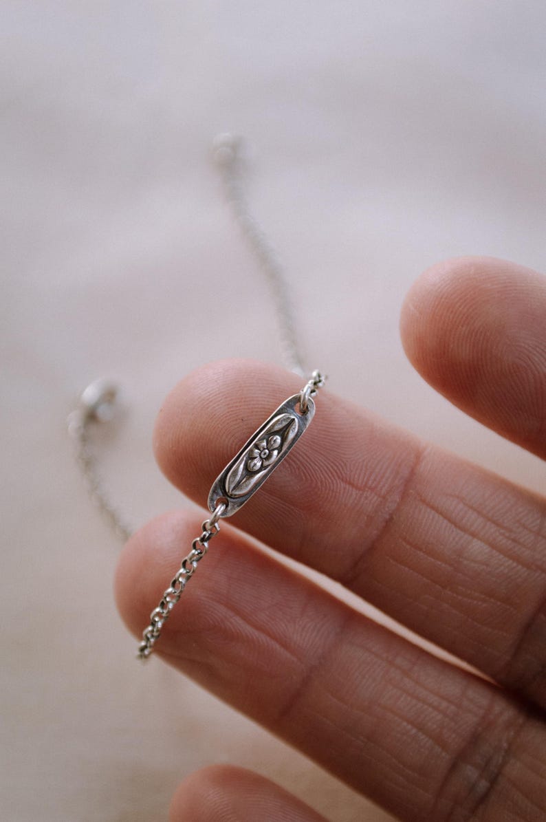 May include: A delicate silver chain bracelet with a small oval charm featuring a floral design.