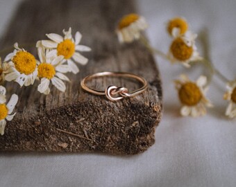 May include: A rose gold knot ring sits on a piece of weathered wood, with dried daisies. The ring features a knot design. The daisies have white petals and yellow centres. The ring is a simple, elegant design.