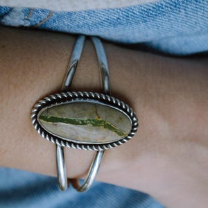 May include: Silver cuff bracelet featuring an oval stone with green and tan hues. The stone is set in a twisted rope design and framed by a silver border. The bracelet is a stylish accessory.