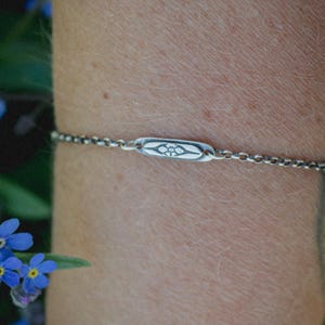 May include: A silver-toned chain bracelet featuring a central, elongated rectangular metal charm. The charm is engraved with a small, symmetrical floral motif centered within a pointed oval outline, connected to a delicate, dark-toned metal link chain.