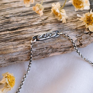 May include: A silver chain bracelet with a rectangular charm featuring a floral design. The bracelet is lying on a piece of weathered wood with dried flowers.