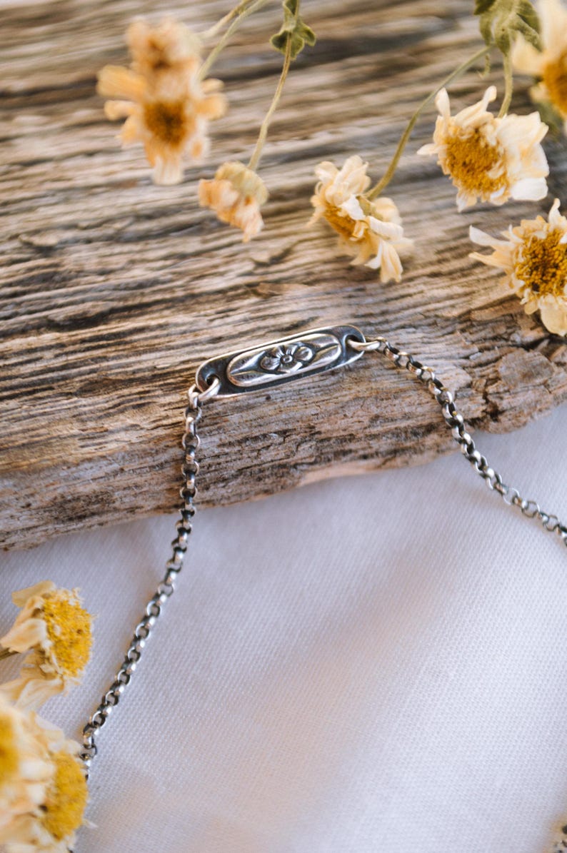 May include: A silver chain bracelet with a rectangular charm featuring a floral design. The bracelet is lying on a piece of weathered wood with dried flowers.