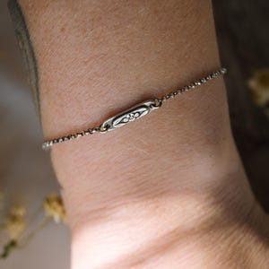 May include: A delicate silver chain bracelet with a small, rectangular charm featuring a floral design.