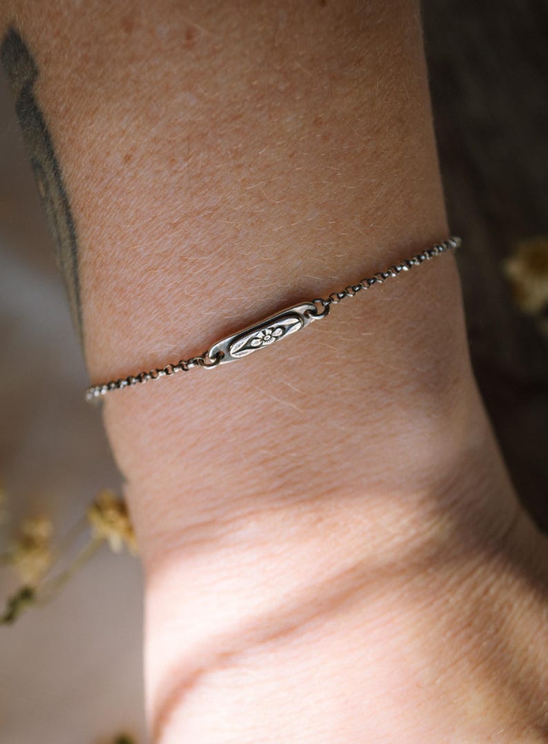 May include: A delicate silver chain bracelet with a small, rectangular charm featuring a floral design.