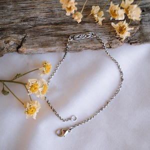 May include: A silver chain bracelet with a small, rectangular charm featuring a floral design. The bracelet is lying on a white fabric surface with dried flowers.