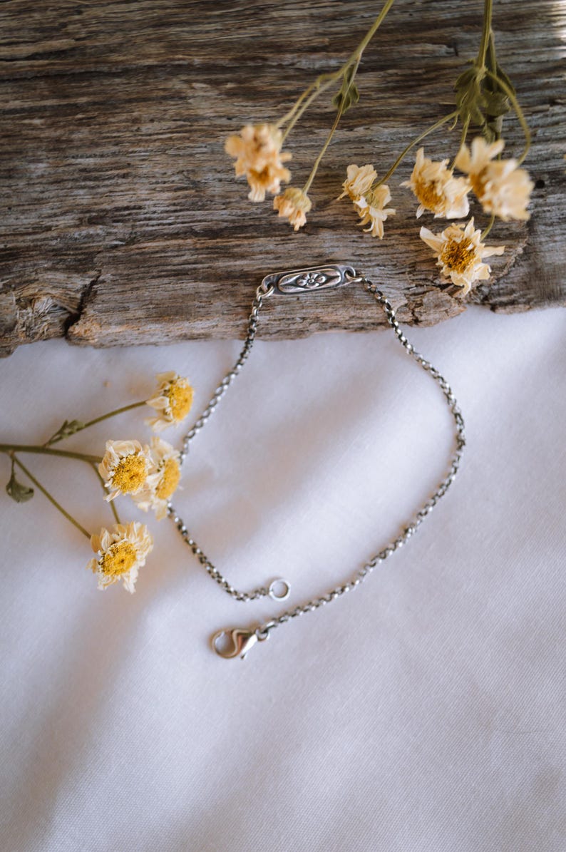 May include: A silver chain bracelet with a small, rectangular charm featuring a floral design. The bracelet is lying on a white fabric surface with dried flowers.