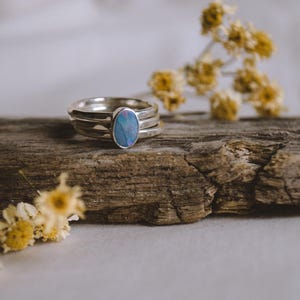 May include: A silver ring with an oval, blue opal stone. The ring is set on a piece of weathered wood, with dried yellow flowers in the background. The ring has a stacked band design.
