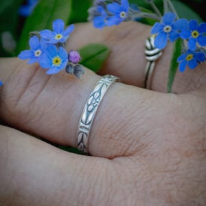 May include: A silver ring featuring an engraved floral pattern on the band, worn alongside a second silver ring with a twisted knot design, positioned among small blue forget-me-not flowers with yellow centers and green leaves.