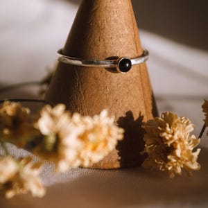 May include: A silver ring with a hammered texture and a black gemstone set in the center. The ring is displayed on a wooden ring holder.