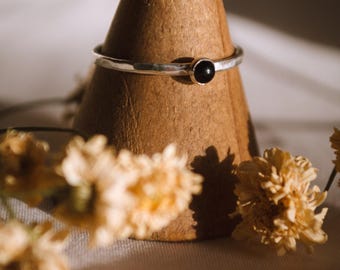 Peut inclure: Une bague en argent avec une texture martelée et une pierre précieuse noire sertie au centre. La bague est présentée sur un porte-bague en bois.
