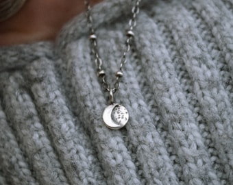 May include: A silver-toned necklace featuring a small, circular pendant with a crescent moon and textured surface detail. The chain is composed of delicate links interspersed with small, round metal beads, creating a subtle, refined appearance.
