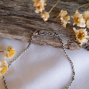 May include: A silver chain bracelet with a small rectangular charm featuring a floral design. The bracelet is lying on a white surface with dried flowers in the background.