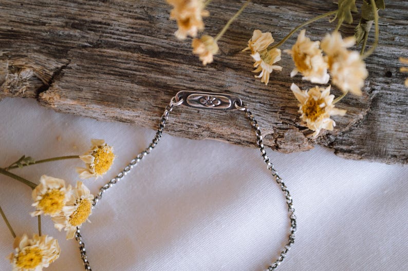 May include: A silver chain bracelet with a small rectangular charm featuring a floral design. The bracelet is lying on a white surface with dried flowers in the background.