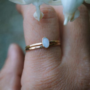 May include: A delicate gold ring with a white opal gemstone. The ring is worn on a finger and is set against a background of white flowers.