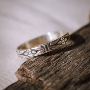 Puede incluir: Un anillo de plata con un diseño floral. El anillo está sobre una pieza de madera desgastada.