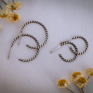 May include: A pair of silver hoop earrings with a twisted design. The earrings are on a white background.