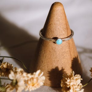 Op de afbeelding: Een delicate zilveren ring met een turquoise edelsteen op een houten ringstandaard. De ring is omringd door gedroogde bloemen.