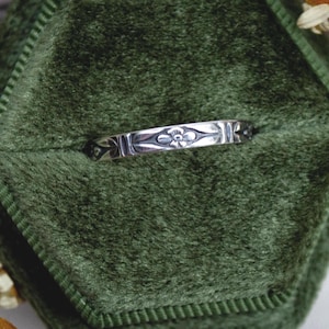 Könnte beinhalten: Ein silberner Bandring mit einem sich wiederholenden, eingravierten Blumenmuster auf der Außenfläche. Das Metall hat eine polierte Oberfläche mit abgedunkelten vertieften Bereichen, die die Details der geätzten Blumenmotive hervorheben.