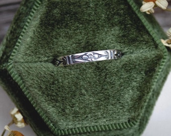 May include: A silver band ring featuring a repeating engraved floral pattern along the outer surface. The metal has a polished finish with darkened recessed areas that highlight the intricate details of the etched flower motifs.