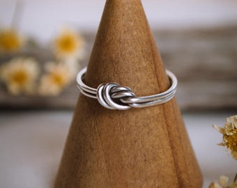 Op de afbeelding: Een zilveren ring met een geknoopt ontwerp, getoond op een houten kegel.