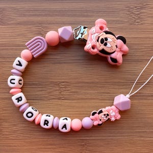 Peut inclure: Un attache-tétine rose et lavande avec le nom "AURORA" épelé en perles de lettres blanches. Le clip présente un charm Minnie Mouse et une perle en forme d'arc-en-ciel. Le clip est attaché à une ficelle blanche.