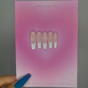May include: Five artificial nails with a white tip and a snowflake design, presented on a pink card. The text "VIRGO NAILS TR" is at the top, and "ARE BEAUTIFUL TO THE TIP OF YOUR NAILS" is at the bottom.