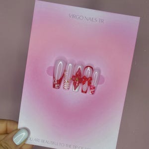 Könnte beinhalten: Ein Set aus langen, Sarg-förmigen Press-On-Nägeln mit einem rosa und roten Design. Die Nägel haben einen Farbverlauf, rote Schleifen und goldene Akzente. Auf der Karte hinter den Nägeln steht "VIRGO NAILS TR" und "YOU ARE BEAUTIFUL TO THE TIP OF YOUR NAILS."