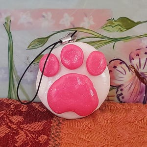 以下が含まれることがあります： ピンクの足跡のデザインが施された、丸い白いチャーム。 足跡はキラキラとした質感で、4つの小さな円と1つの大きな円で構成されています。 黒いコードとシルバーの留め金がチャームに付いています。