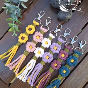 Peut inclure: Plusieurs porte-clés en macramé faits à la main, ornés de motifs floraux colorés. Chaque porte-clés est doté d'un fermoir argenté et d'une frange en fil assorti. Les fleurs sont dans les tons de jaune, rose, blanc, violet et bleu, avec des tiges vertes. Le texte "Cordellettes & Cacahuètes" est en arrière-plan.