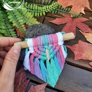 Peut inclure: Une petite suspension murale en macramé avec une baguette en bois, présentant un arc-en-ciel de couleurs, notamment le rose, le violet, le bleu et le vert. Un charme en plume argentée pend du centre du macramé.