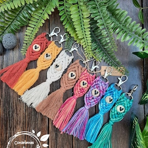 Peut inclure: Un ensemble de huit porte-clés en macramé colorés avec des breloques en forme de cœur en bois. Chaque porte-clés présente une combinaison de couleurs de fil différente, notamment orange, jaune, blanc, marron, rose, violet, bleu et vert. Les porte-clés sont faits à la main et ont un pompon en bas.