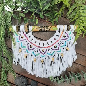 Peut inclure: Suspension murale en macramé blanc avec une baguette en bois et des perles colorées. Le macramé a la forme d'un demi-cercle et est doté d'une frange en bas. La baguette en bois présente un motif géométrique noir et blanc.