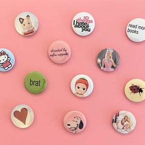 May include: A collection of colorful round buttons with various designs and text. The buttons feature images of a cartoon cat, a fairy, a ladybug, and Snoopy. Some buttons have text that reads "10 things I hate about you", "read more books", and "brat".