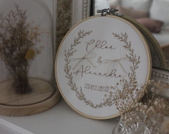 May include: A circular wooden embroidery hoop containing white fabric with the embroidered names Chloe & Alexandre, a date 23.02.2023, and a surrounding botanical wreath design. Two thin beige ribbons are tied in bows on the fabric surface.