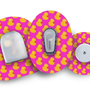 Op de afbeelding: Drie ronde medische apparaatpleisters met een roze achtergrond en een geel rubberen eendjespatroon. De pleisters zijn voor een continue glucosemeter.